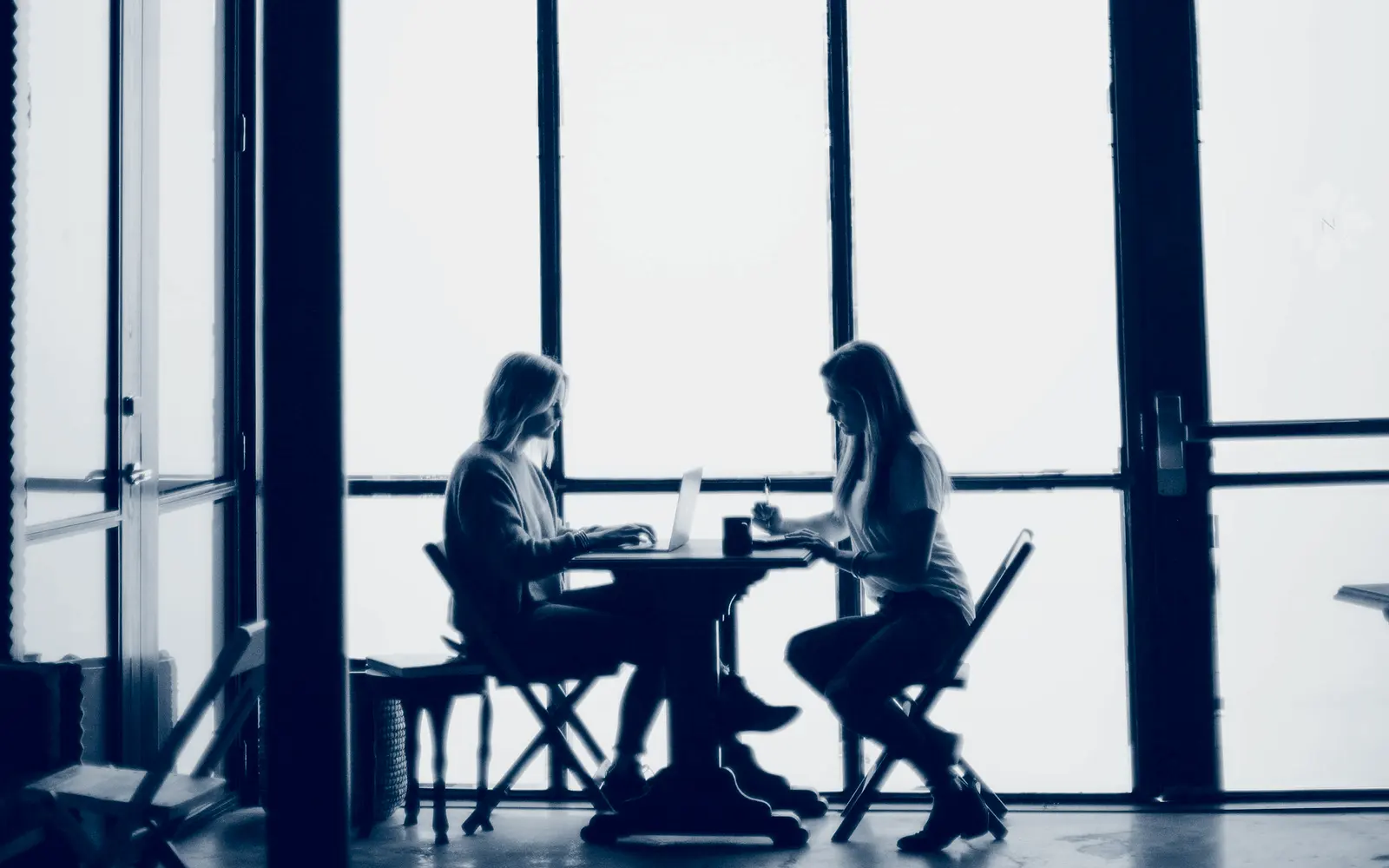 Twee mensen in gesprek aan een tafel bij een groot raam.